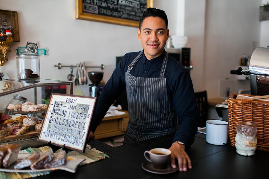Business Insurance - Cafe Owner Smiles Behind the Counter Wearing a Gray Apron